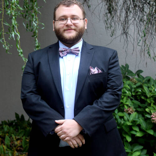 louis Marx is wearing a dark blue suit, white shirt, and a patterned bow tie with a matching pocket square, standing with hands clasped in front of a green leafy background